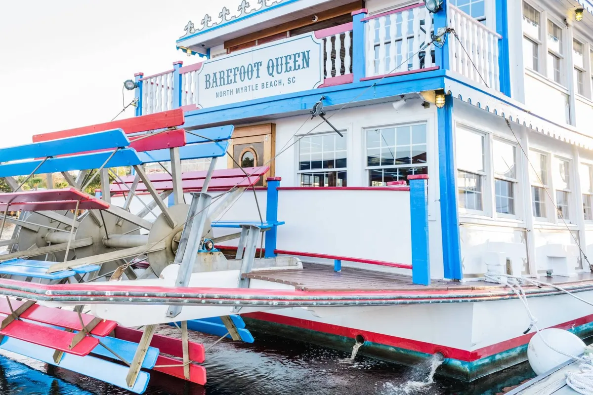 The vibrant paddle wheel of the Barefoot Queen Riverboat stands out against the colorful exterior and clear signage. The boat is docked and ready for guests, hinting at lively cruises, dining, and entertainment on the water.