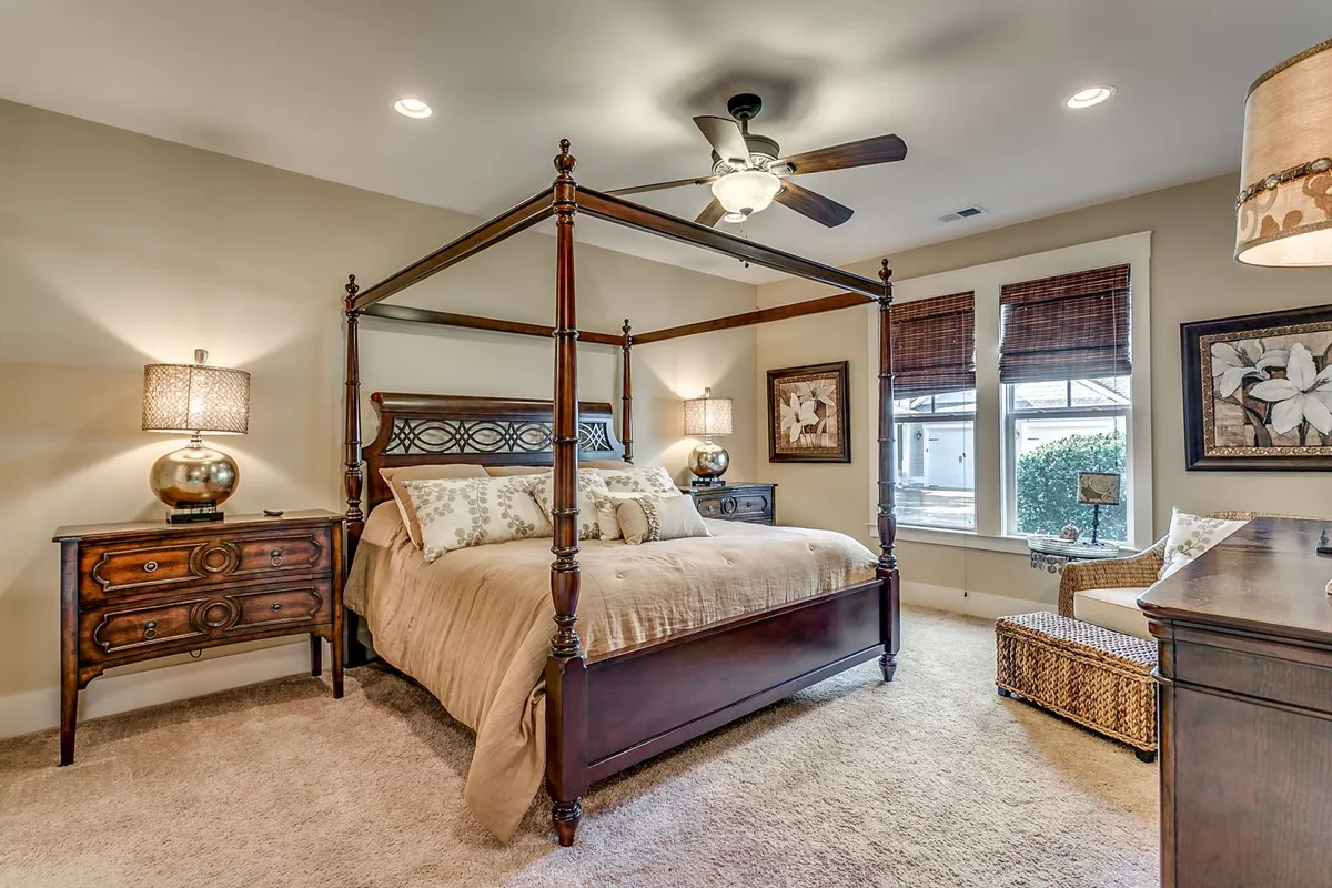 A serene bedroom featuring a four-poster bed with crisp linens, decorative lamps flanking the bedside, a window with blinds softly filtering natural light, and tasteful artwork adorning the walls. The atmosphere is tranquil and restful, ideal for relaxation and reading.