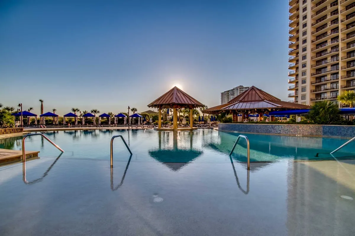 A luxurious pool scene at sunset featuring a large swimming pool, thatched-roof cabanas, and a relaxed, upscale mood. The golden light enhances the resort ambiance, suggesting an ideal spot for evening relaxation and socializing by the water.