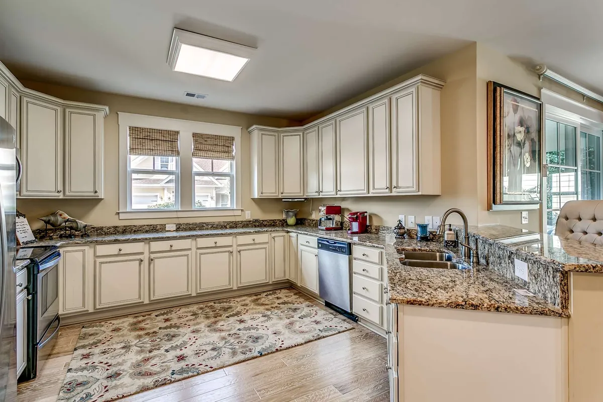A modern, fully-equipped kitchen with granite countertops, stainless steel appliances, a central kitchen island, and stylish window treatments. A decorative rug adds warmth, and the space is clearly designed for both cooking and socializing.