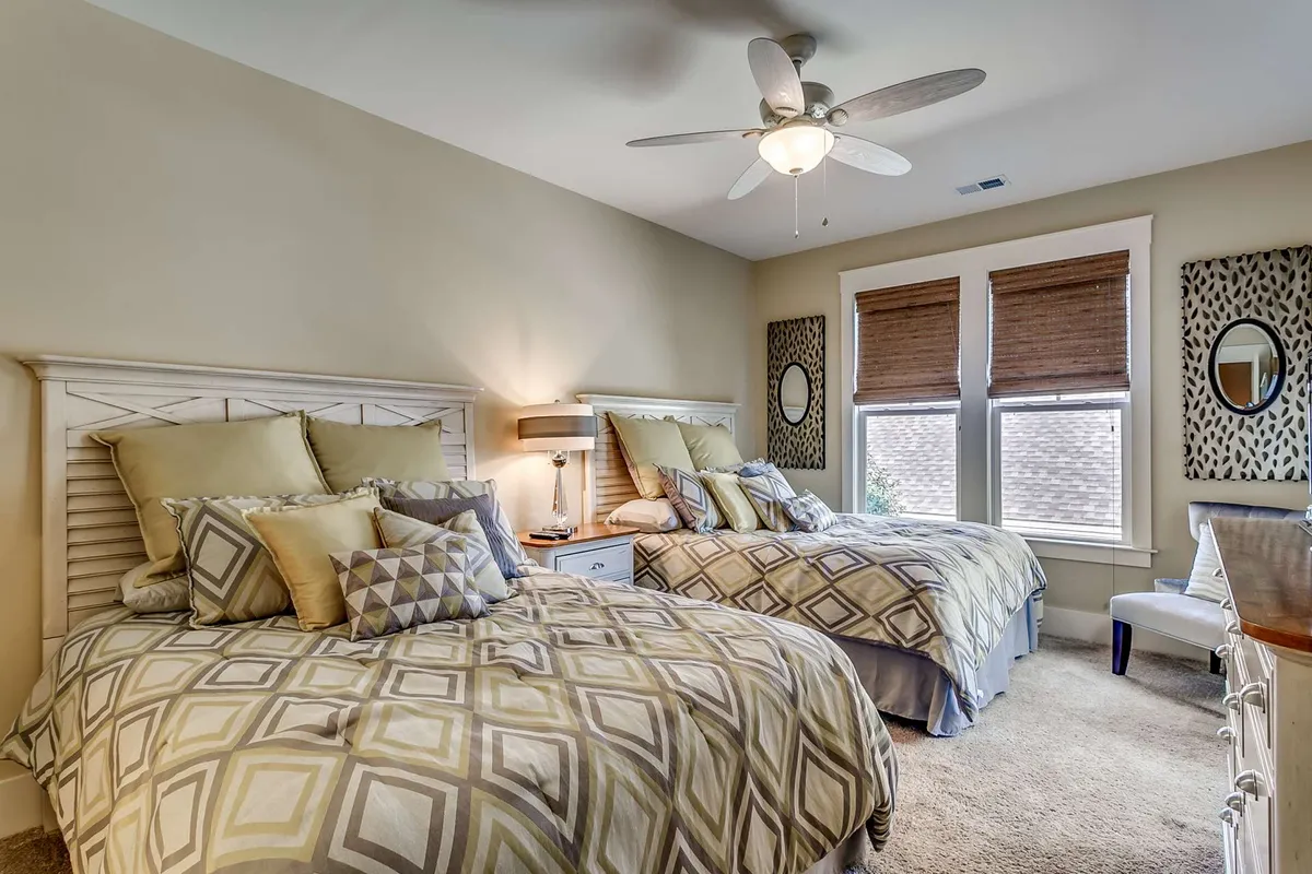A spacious bedroom with two queen-sized beds adorned with decorative pillows, ceiling fan overhead, and large windows with blinds that let in gentle daylight. The room feels inviting and is ideal for families or groups, providing ample sleeping space and comfort.