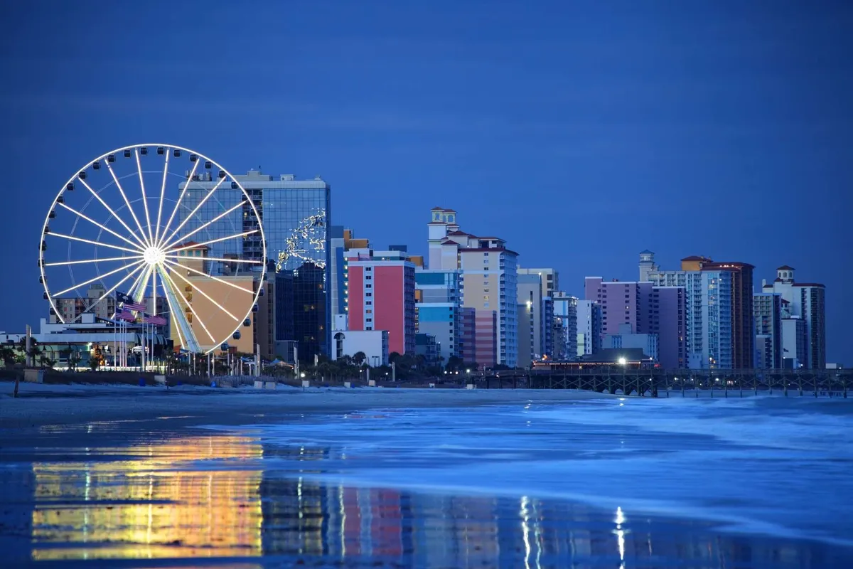 A lively scene with a colorful ferris wheel, beachfront buildings, and ocean waves. The area is bustling with people enjoying carnival rides, walking along the beach, and visiting nearby restaurants, all under a festive and cheerful atmosphere.