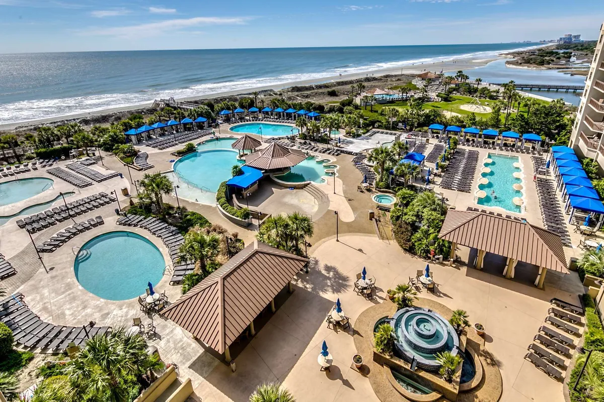 This exterior image showcases a sprawling pool complex with several swimming pools, an ocean view in the background, landscaped gardens, and a cabana area. Guests can be imagined swimming, sunbathing, and relaxing in a tranquil, resort-like environment.