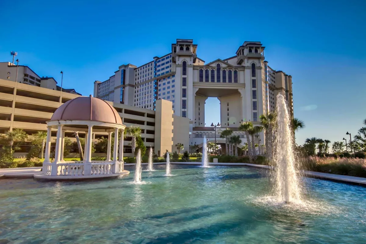 A luxurious outdoor setting featuring an ornate gazebo surrounded by lush gardens, shooting fountain water jets, and grand hotel architecture. The scene is inviting, perfect for relaxing or group photos in a resort atmosphere.